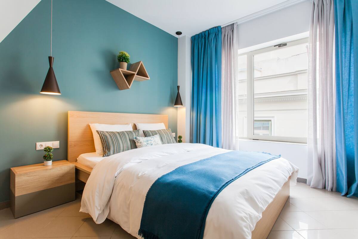 A modern bedroom with a bed, bedside tables, a hanging plant, and a window with curtains.
