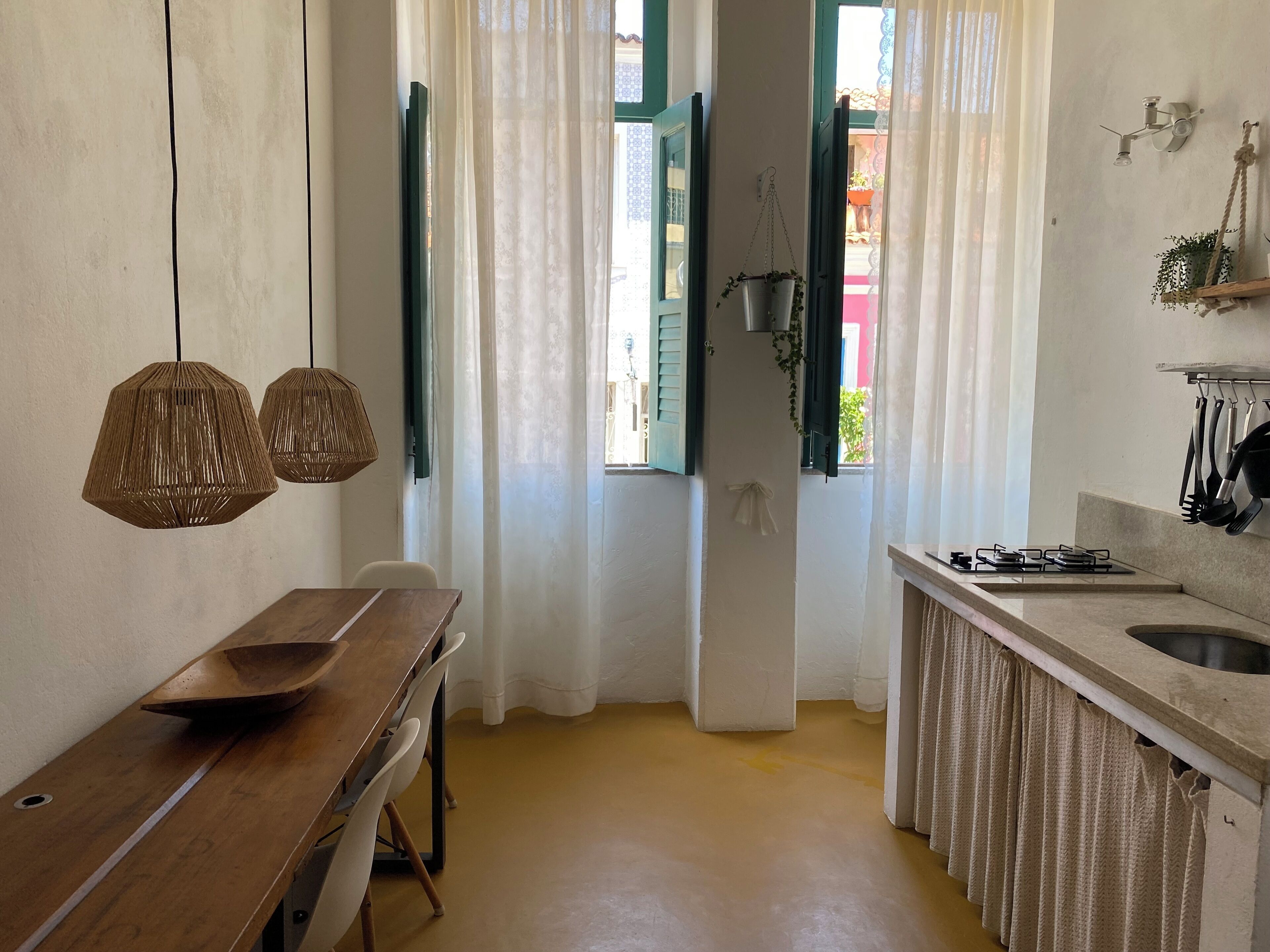 Una cocina con una mesa de madera, dos lámparas colgantes, un ventanal con cortinas y una planta suspendida.