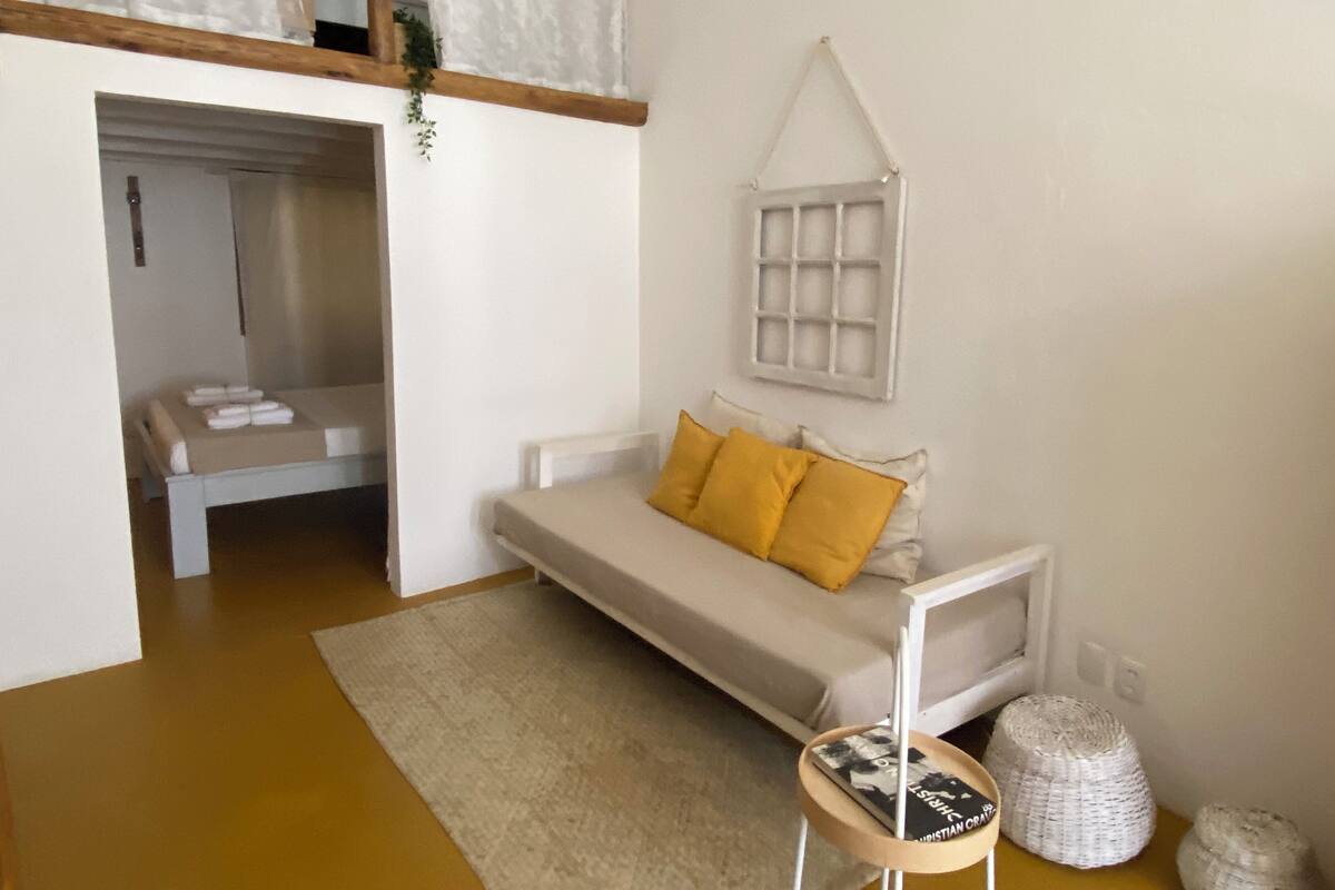 A modern, minimalist bedroom with a bed, a small seating area, and a hanging light fixture.