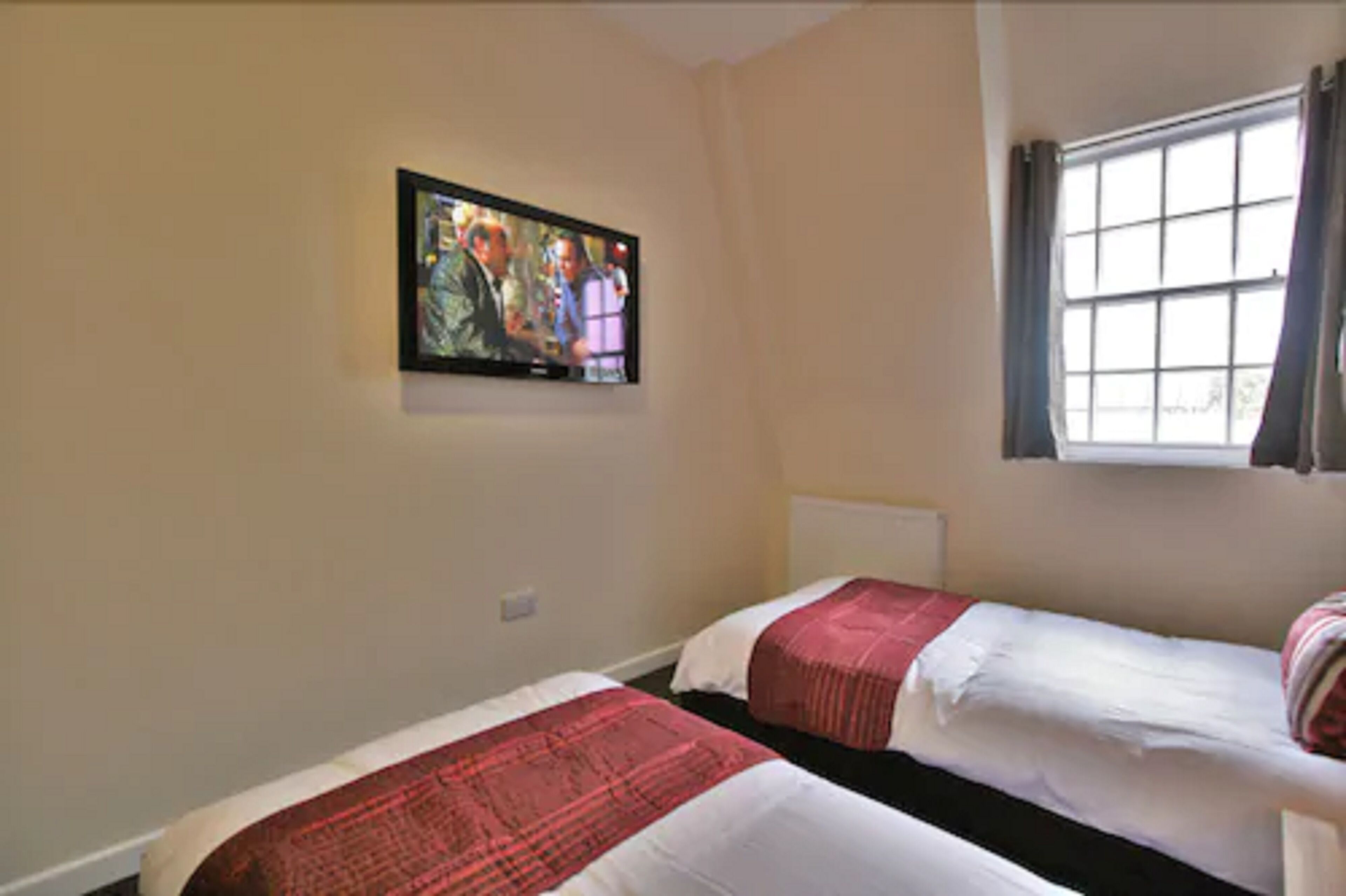 A room with two beds, a TV mounted on the wall, and a window with curtains.
