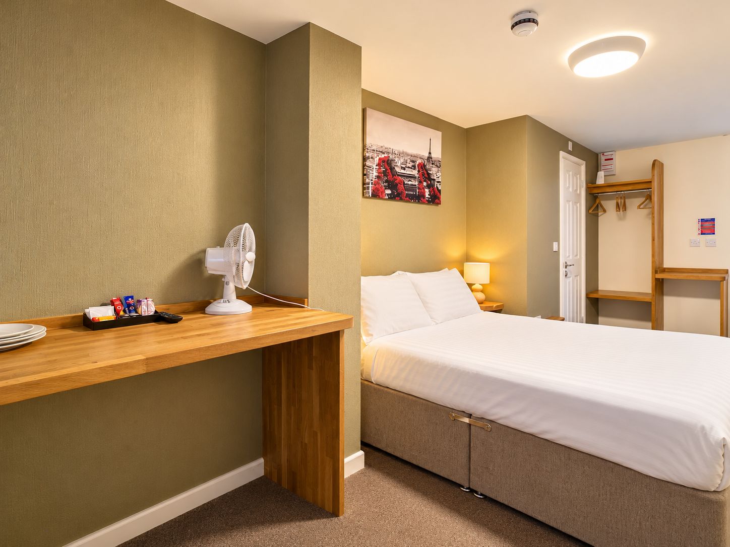 A hotel room with a bed, bedside table, fan, and a wall-mounted picture.