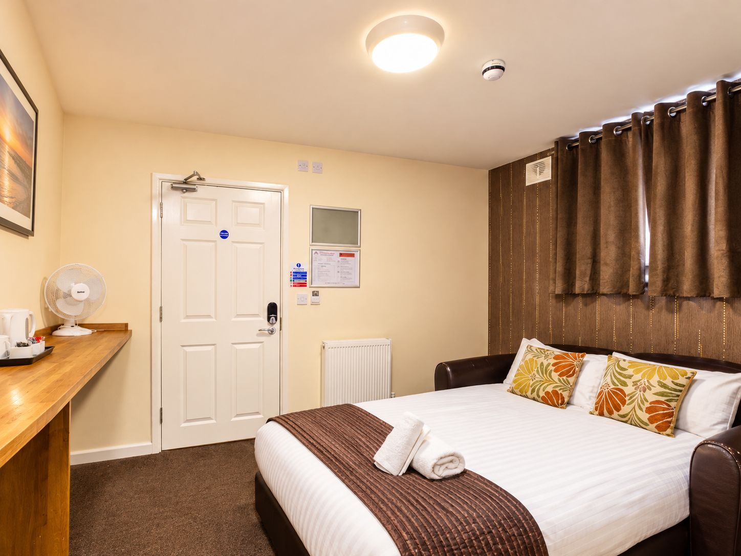 A hotel room with a bed, a desk with a fan, a door, a wall-mounted control panel, and a window with curtains.