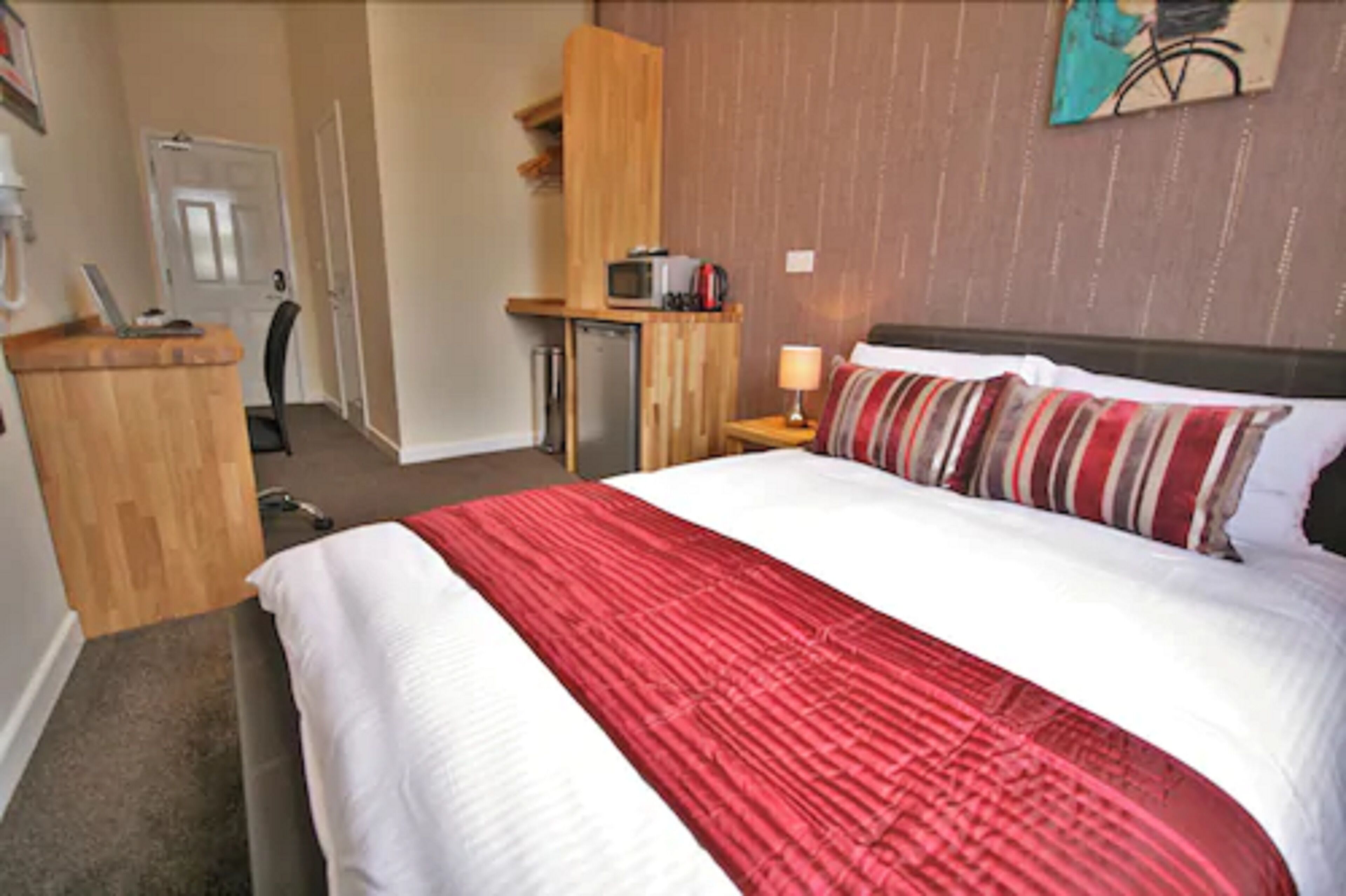 A hotel room with a bed, a desk, a chair, a microwave, a lamp, and a door.