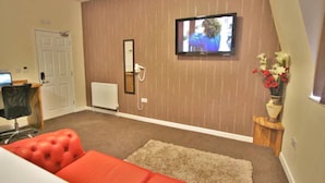 A room with a television mounted on the wall, a red sofa, a small table with a vase of flowers, and a door with a computer on a desk. - Homesly Gloucester Hotels Ltd. (Gloucester)