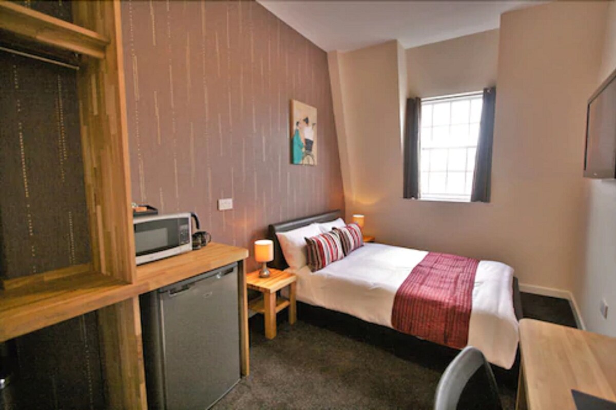 Une chambre d’hôtel compacte comprenant un lit, une table de chevet, une lampe et une petite kitchenette équipée d’un four à micro-ondes.
