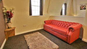 A room with a red leather sofa, a beige carpet, a wooden side table with a vase of flowers, and a window with curtains. - Homesly Gloucester Hotels Ltd. (Gloucester)