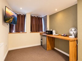 A compact kitchen with a microwave, kettle, and fan, a TV mounted above a window with curtains, and a dishwasher.