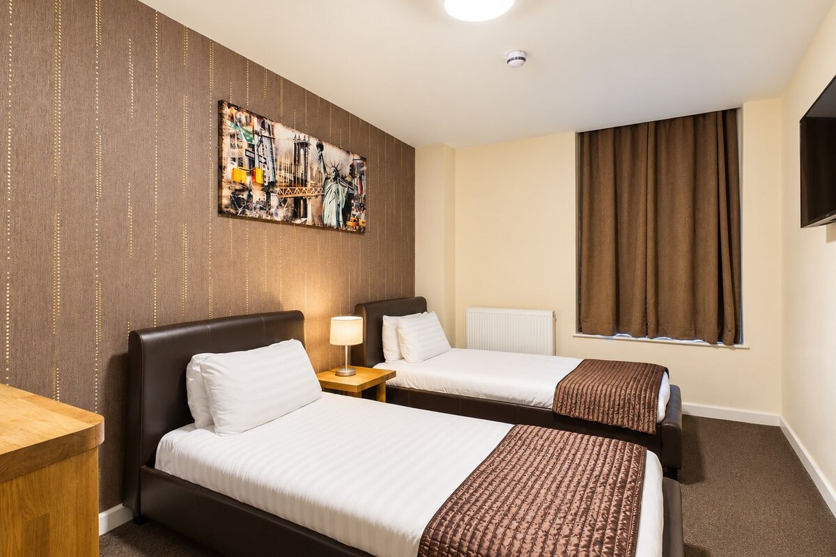 Hotellihuone, jossa on kaksi sänkyä, seinällä taulu, ikkunassa verhot ja seinällä televisio.