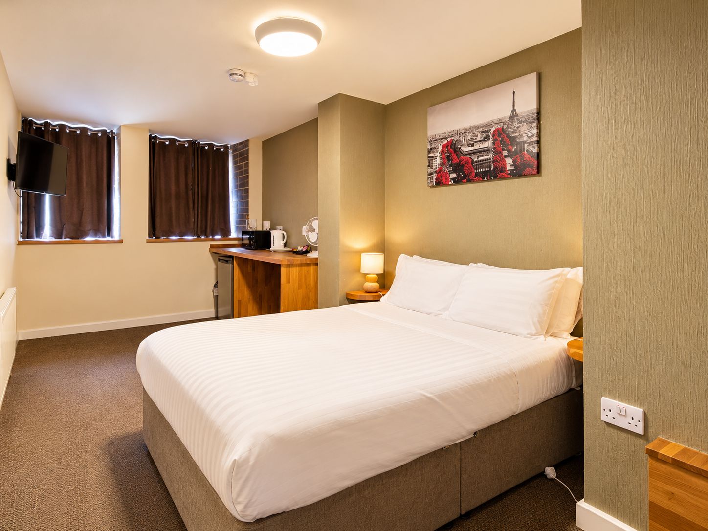 A hotel room with a bed, a desk with a coffee maker, a TV mounted on the wall, and a window with curtains.