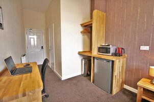 A compact kitchenette with a microwave, coffee maker, and a small refrigerator. There is a laptop on a wooden table, a chair, and a trash bin.