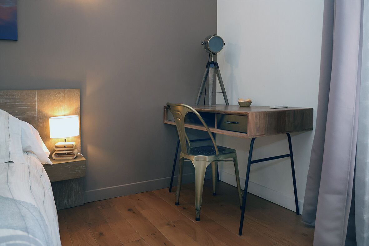 A bedroom with a bed, bedside table with a lamp, a desk with a chair, and a tripod lamp.