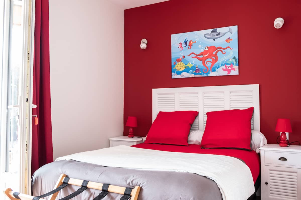 A bedroom with a red wall, a white bed with red and grey bedding, two bedside tables with red lamps, and a window with red curtains.