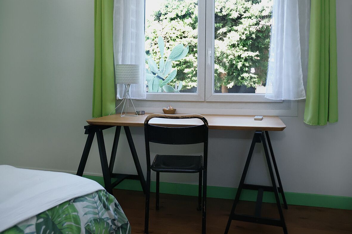 A room with a wooden desk, a black chair, a small table with a lamp, and a window with green curtains.