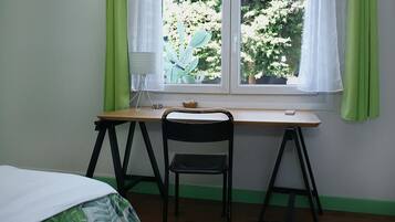 Ein Zimmer mit Holzschreibtisch, schwarzem Stuhl, kleinem Tisch mit Lampe und Fenster mit grünen Vorhängen.