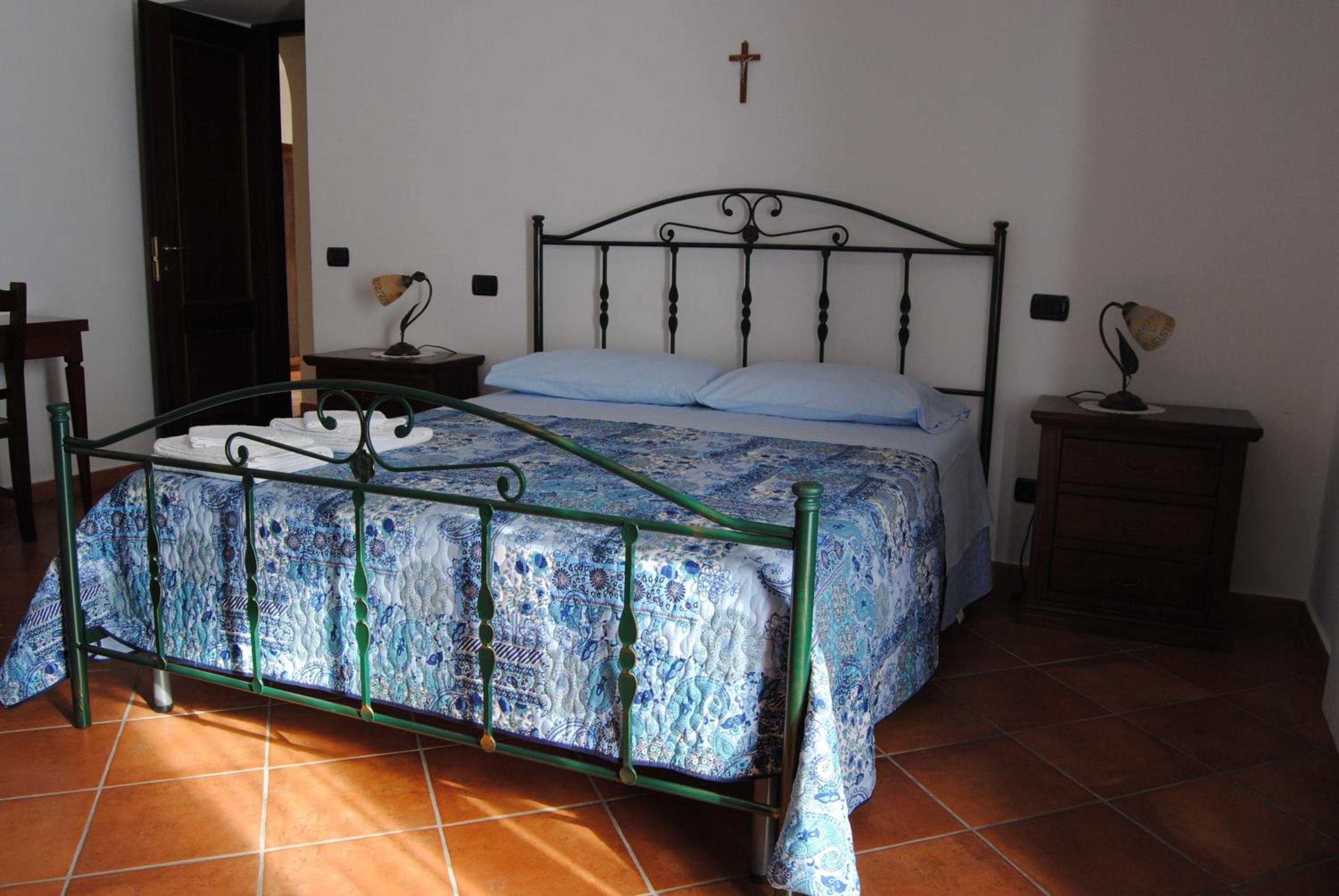 A bedroom with a metal bed frame, a patterned bedspread, two bedside lamps, a wooden nightstand, and a cross on the wall.