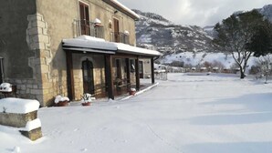Un bâtiment en pierre doté d’un porche abrité, de plantes en pot et d’un paysage enneigé avec des montagnes en arrière-plan.