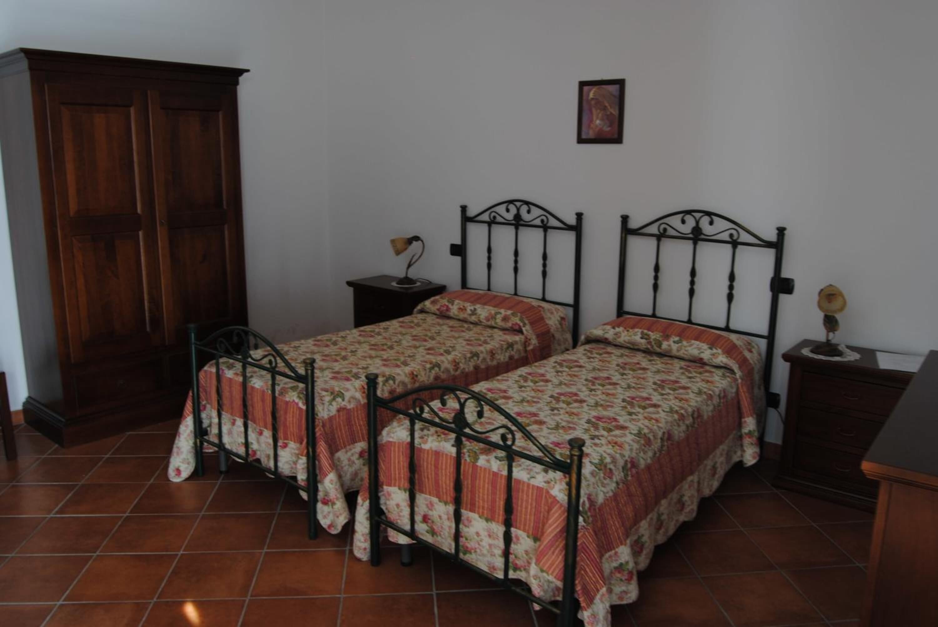 Quarto com duas camas de solteiro, um guarda-roupa de madeira, uma pequena mesa de cabeceira com uma luminária e um quadro na parede.