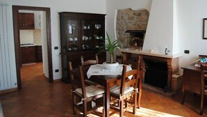 Un coin repas avec une table dressée pour quatre personnes, entourée de chaises en bois. On aperçoit un buffet aux portes vitrées sur la gauche, et une cheminée en pierre se trouve au fond.