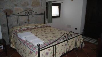 Een slaapkamer met een stenen muur, een bed met een bloemenbedekking, een klein raam met gordijn en een betegelde vloer.