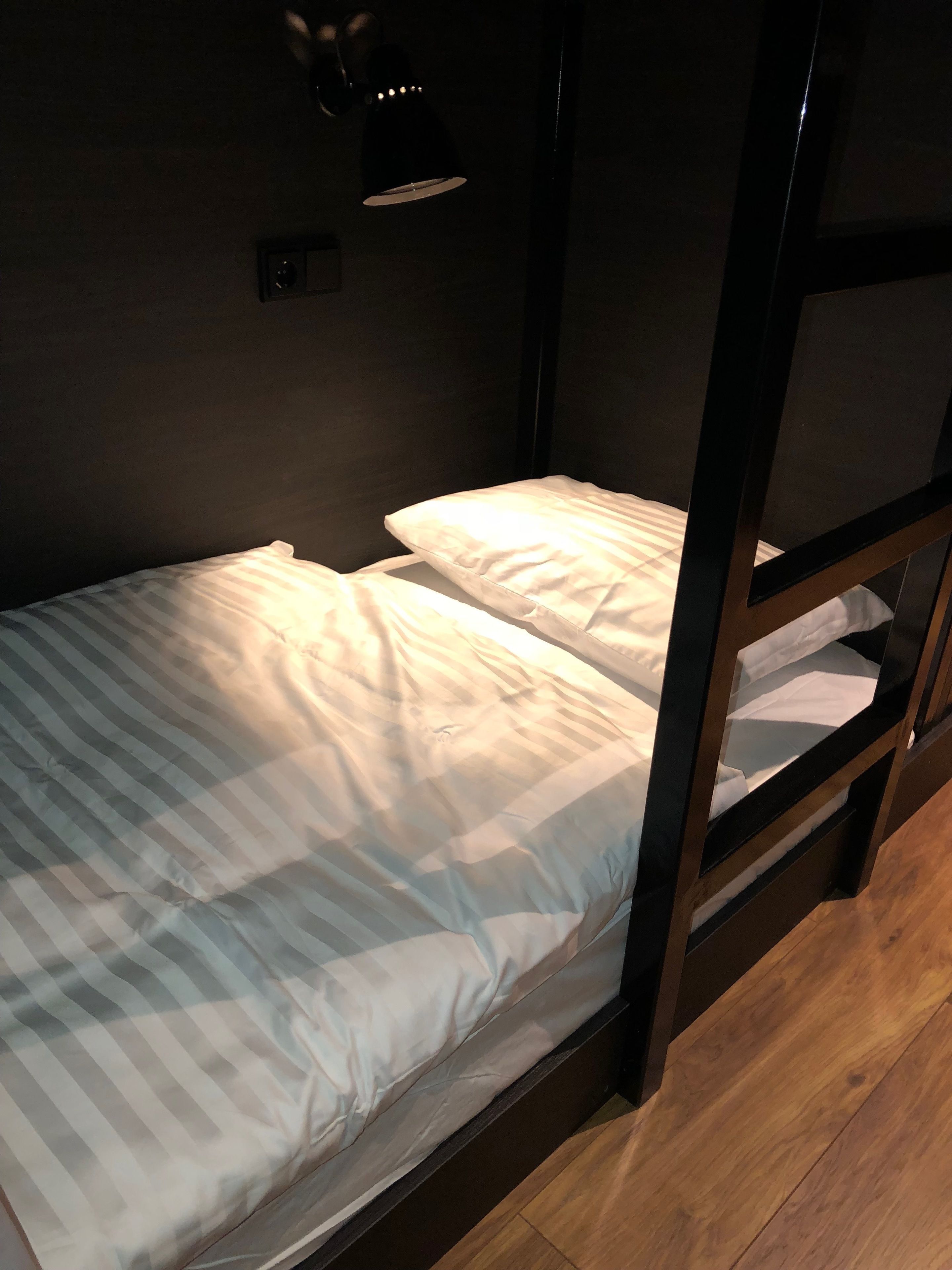A bunk bed with a striped mattress and pillows, a small bedside table with a lamp, and a wooden floor.