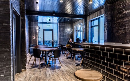 A modern interior with wooden flooring, black brick walls, and large windows offering a city view. Tables and chairs are arranged for dining.