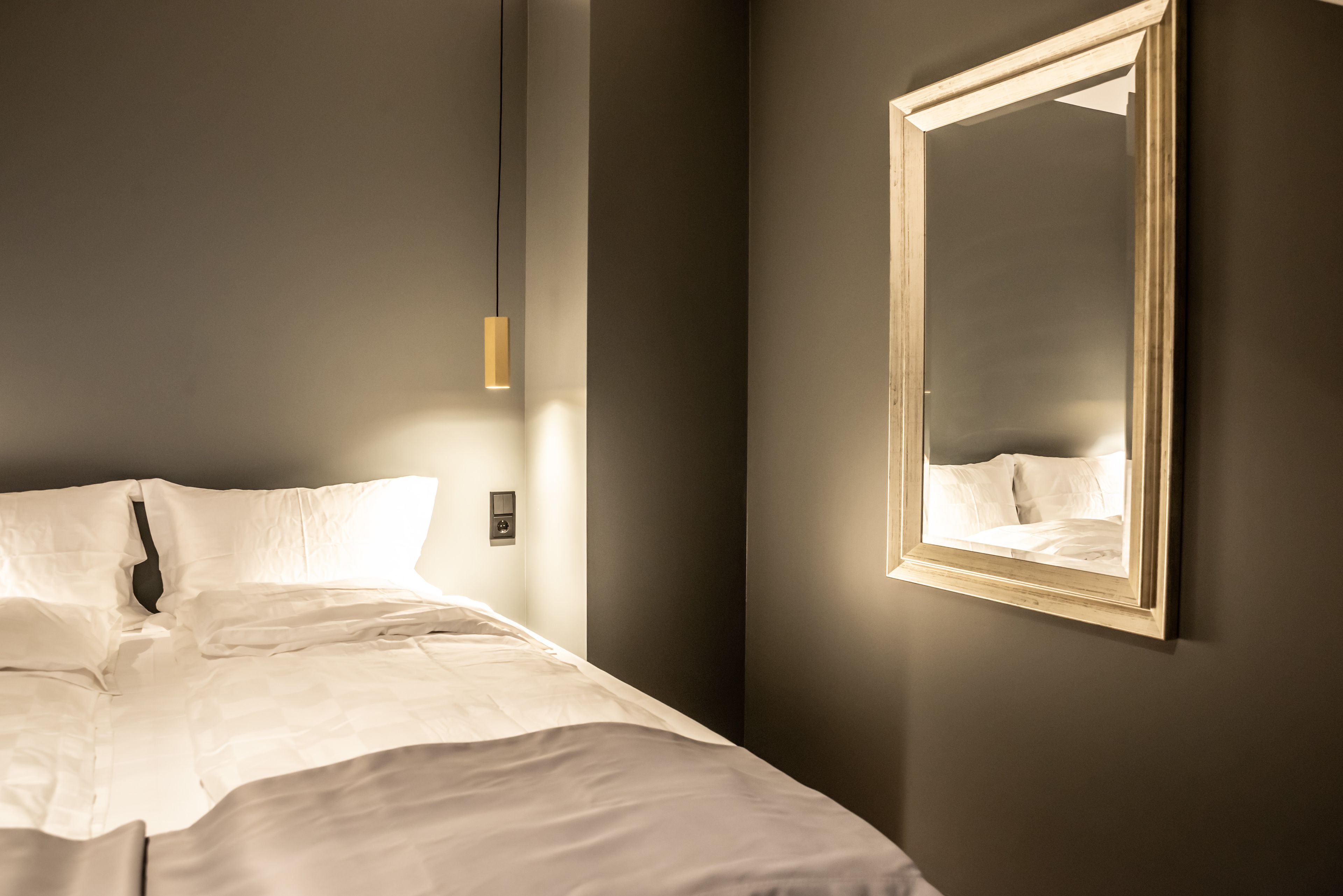 A neatly made bed with white linens, a hanging pendant light, and a mirror on the wall.