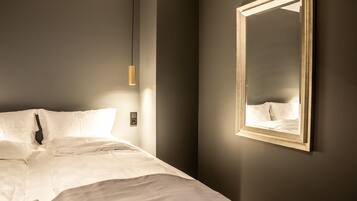 A neatly made bed with white linens, a hanging pendant light, and a mirror on the wall.
