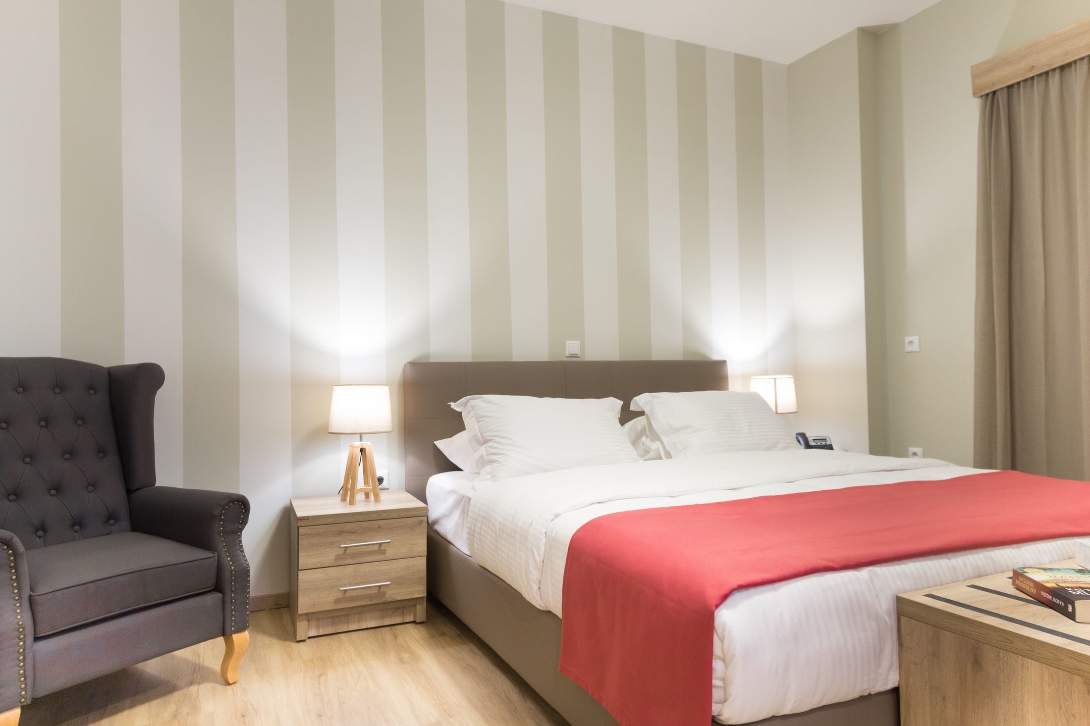 A hotel room with a bed, bedside table, lamp, chair, and a red throw blanket.