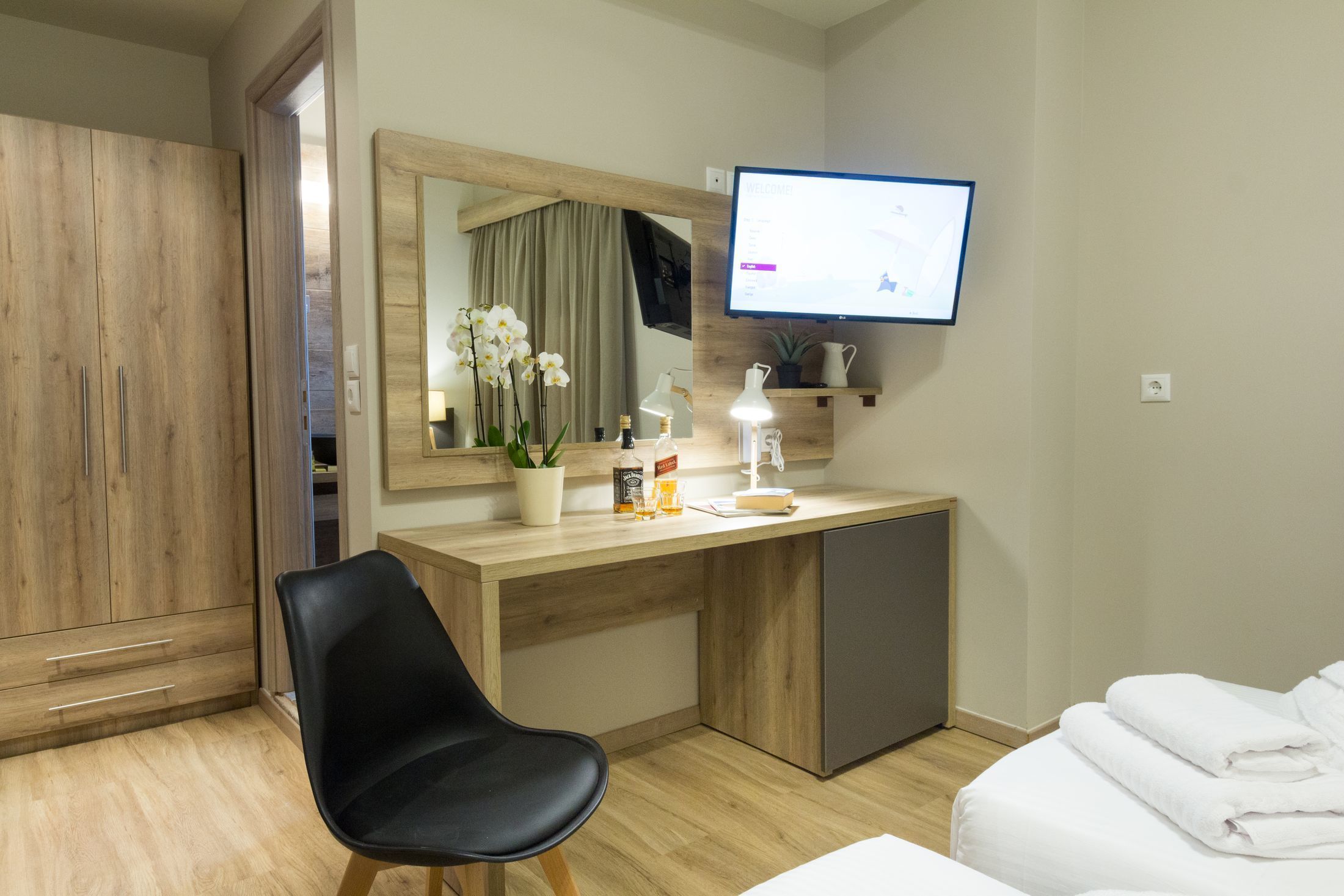 A hotel room with a wooden desk, a black chair, a bed with white linens, a television on the wall, and a potted orchid.