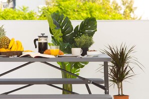 Une table dressée pour le petit-déjeuner, avec un livre, une machine à café, un verre de jus d’orange, un bol de céréales, un ananas, des bananes et un croissant.