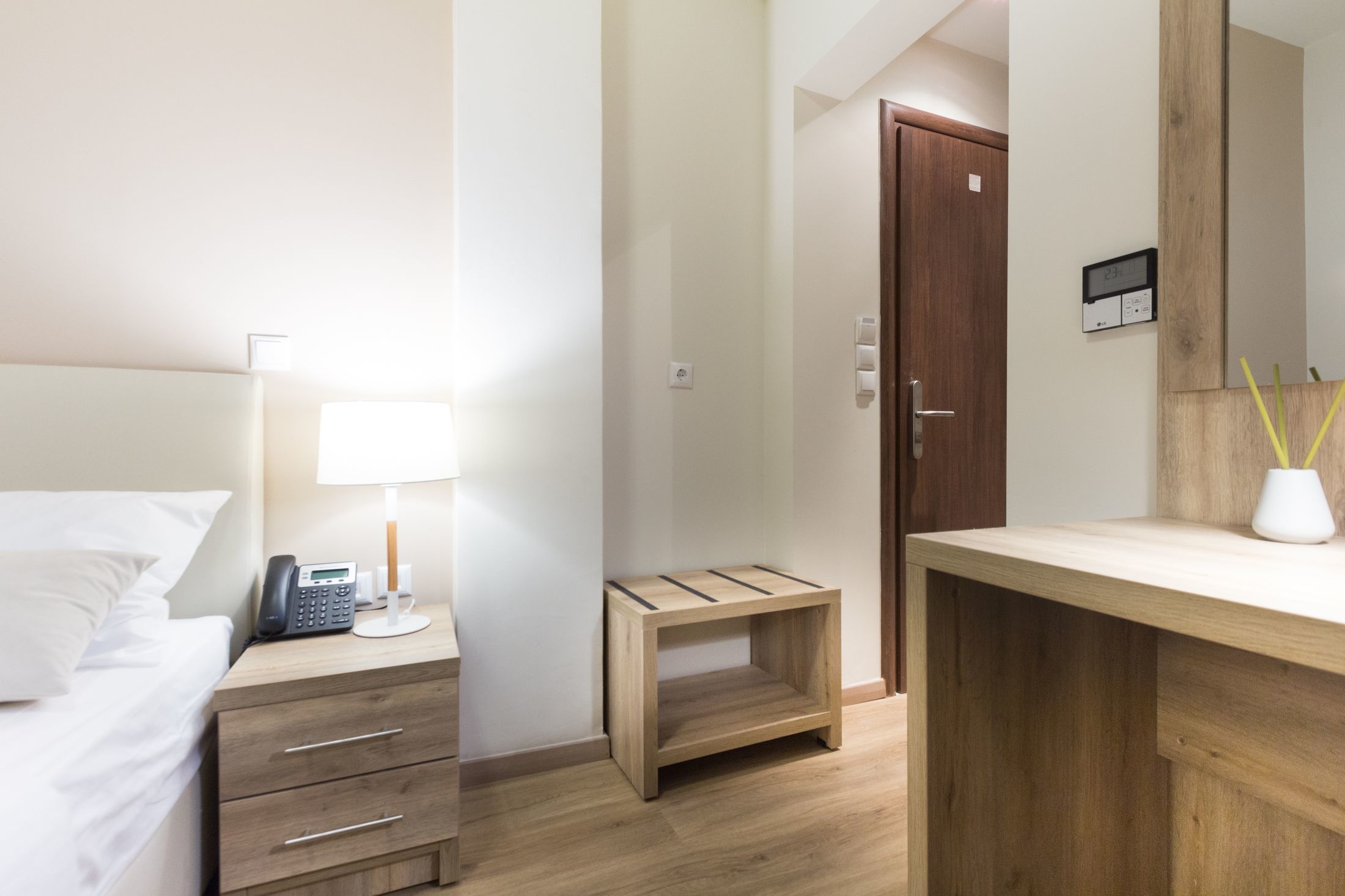 A hotel room with a bed, bedside table with a lamp, a wooden nightstand, a small wooden stool, a wooden desk, and a mirror.