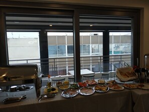 A buffet table with various dishes and a view of the city through large windows.