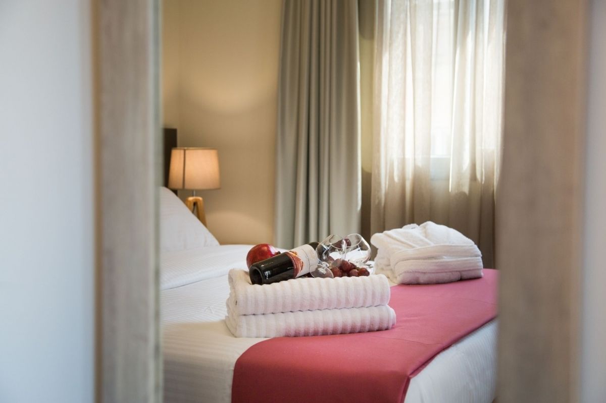 A neatly made bed with white and red bedding, a set of towels, a bottle, and a glass.