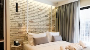 A hotel room with a brick wall, a bed with white linens, a bedside table with a phone, and a window with curtains. - Hotel @ Syngrou Fix (Athens)