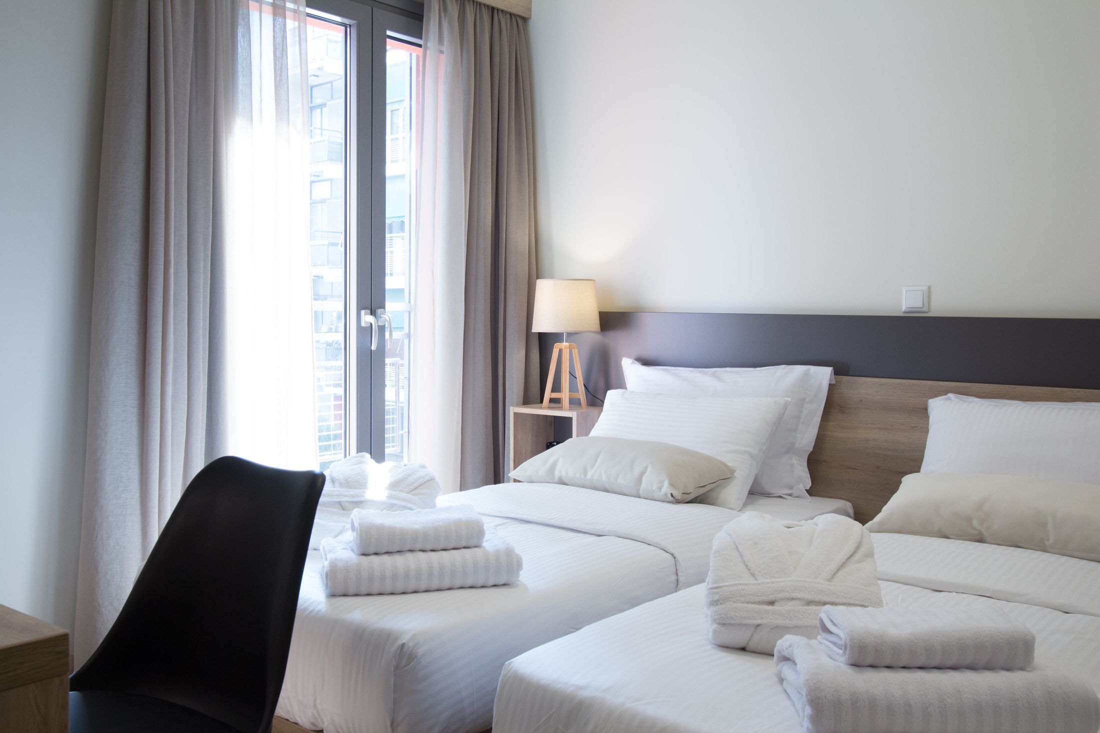 A hotel room with two beds, a chair, a lamp, and a view of the city through the window.
