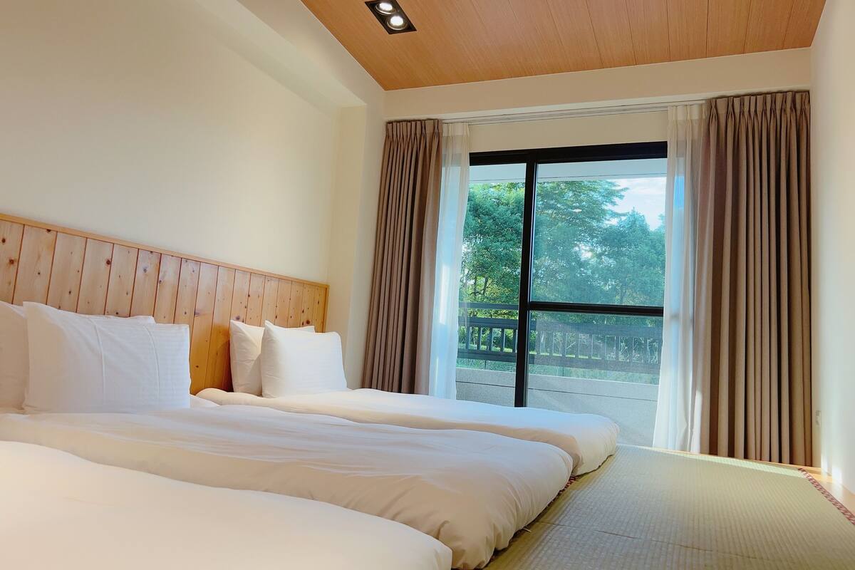 A hotel room with a wooden ceiling, two beds with white bedding, a large window with curtains, and a view of greenery outside.