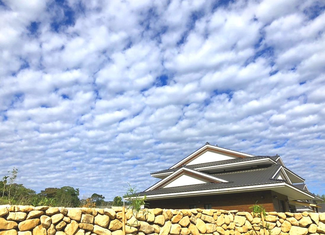 A traditional Japanese-style house with a stone wall in the foreground and a clear blue sky with clouds.