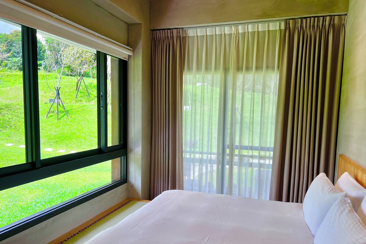 A hotel room with a large bed, a window with curtains, and a view of a grassy area.