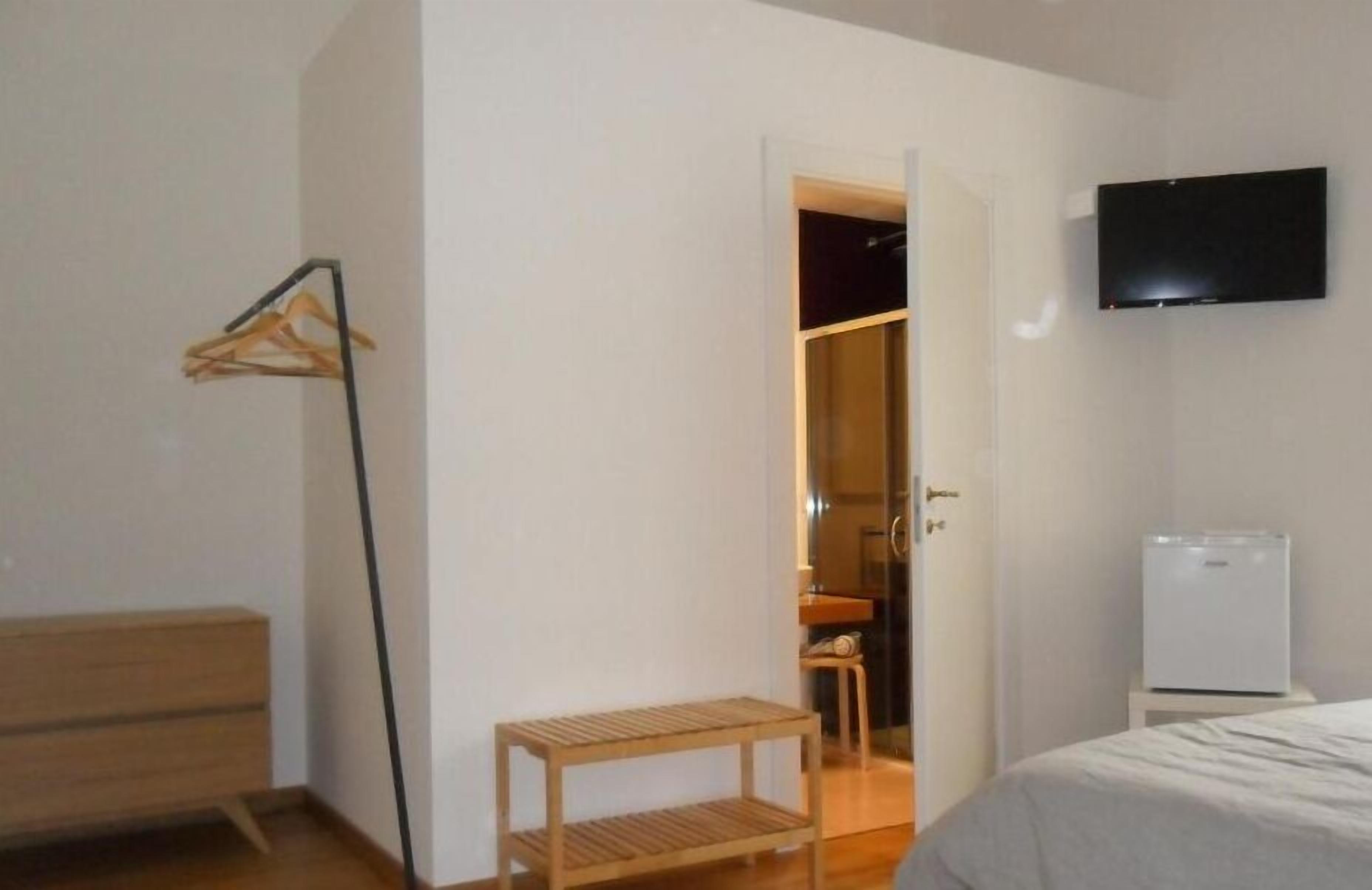Une chambre à coucher comprenant un lit, un banc en bois, un porte-manteau, un téléviseur et un réfrigérateur blanc.