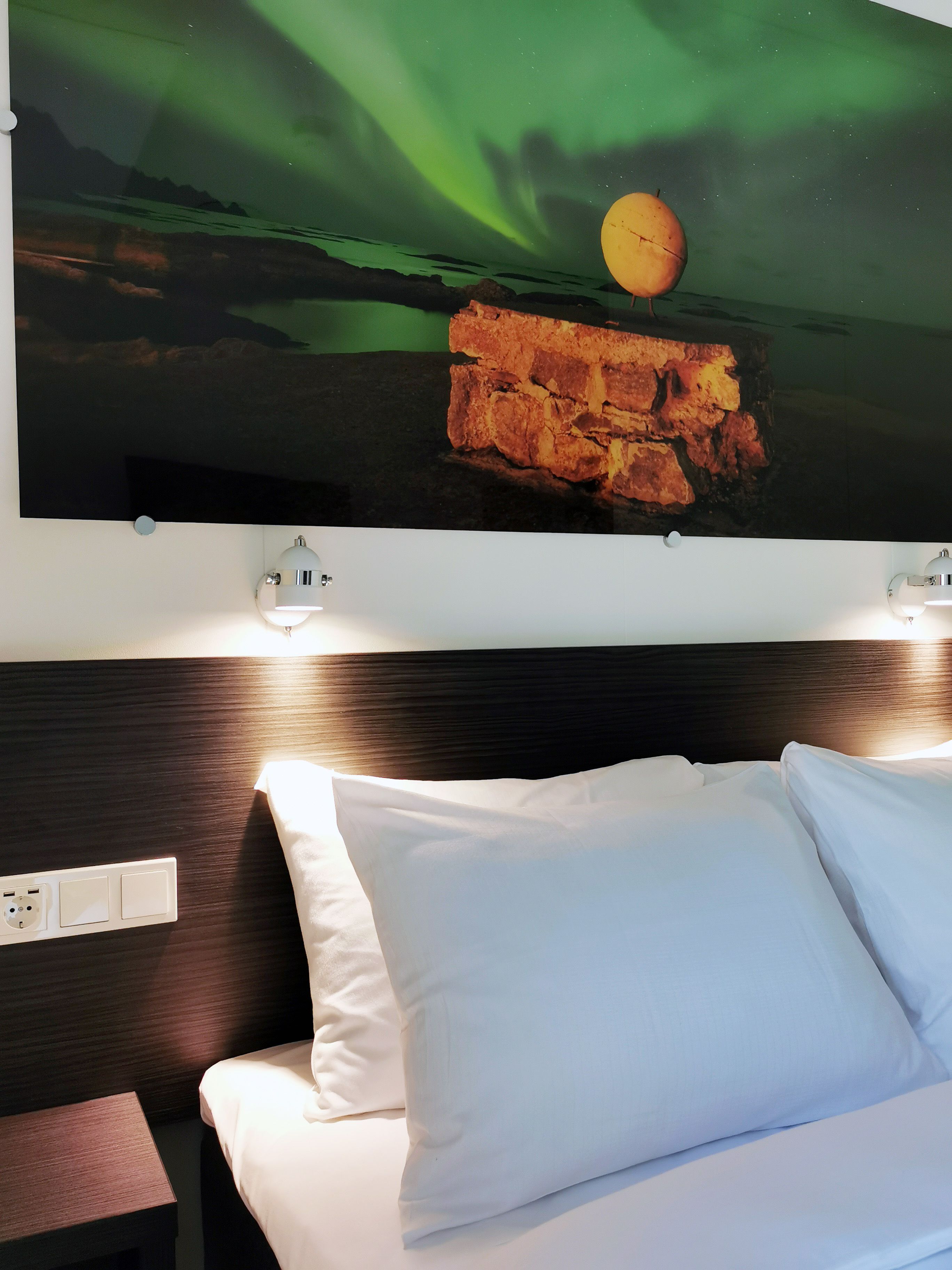 Habitación de hotel con cama, almohadas blancas, cabecera y una obra de arte en la pared que representa un muro de ladrillos y fruta.