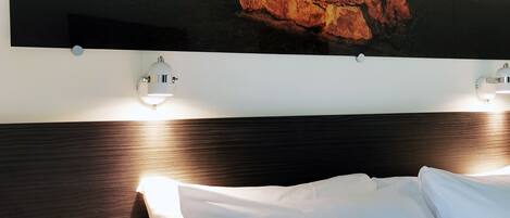 Habitación de hotel con cama, almohadas blancas, cabecera y una obra de arte en la pared que representa un muro de ladrillos y fruta.