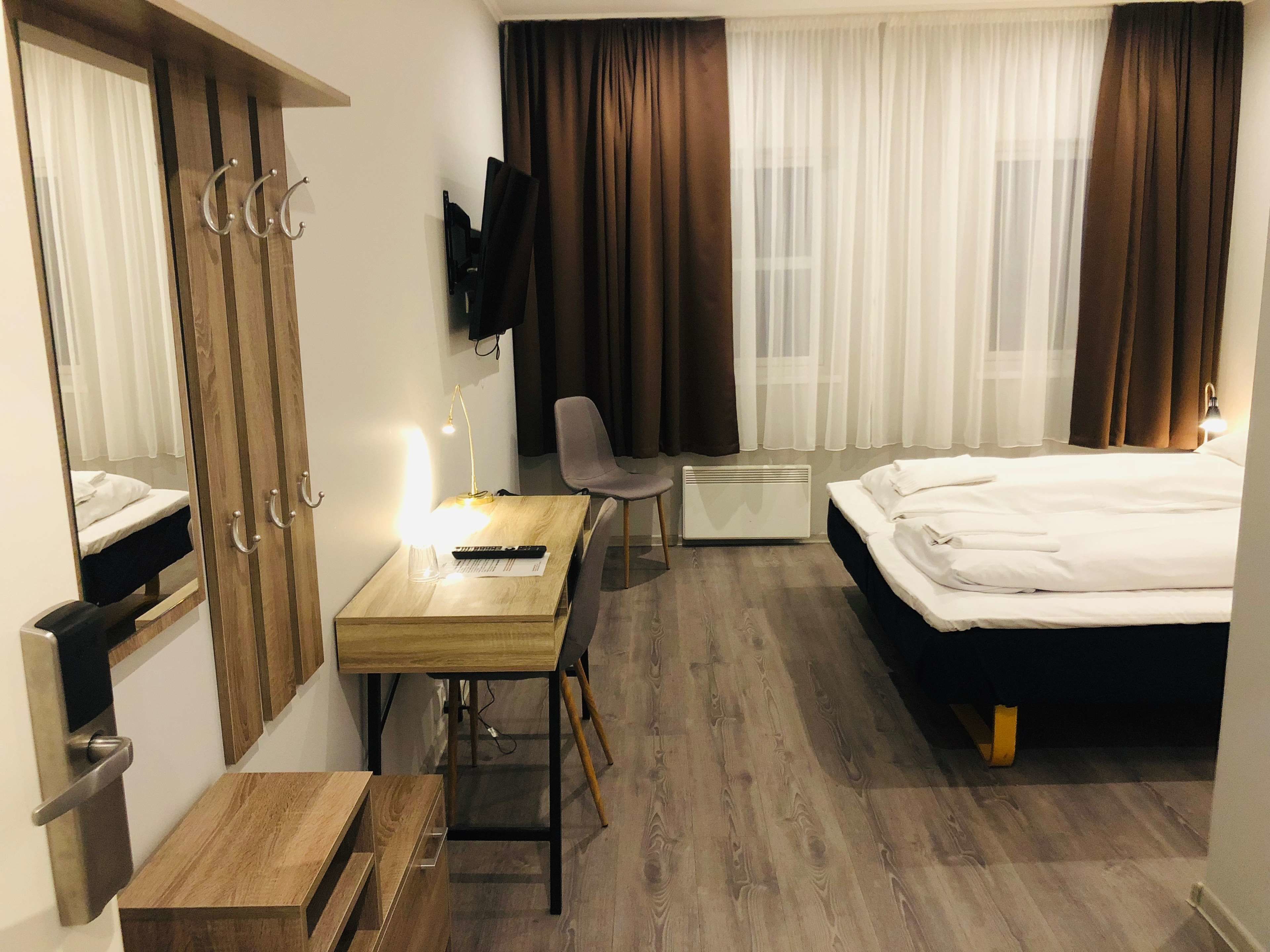 Quarto de hotel com cama, escrivaninha com luminária, cadeira, televisão e guarda-roupa.