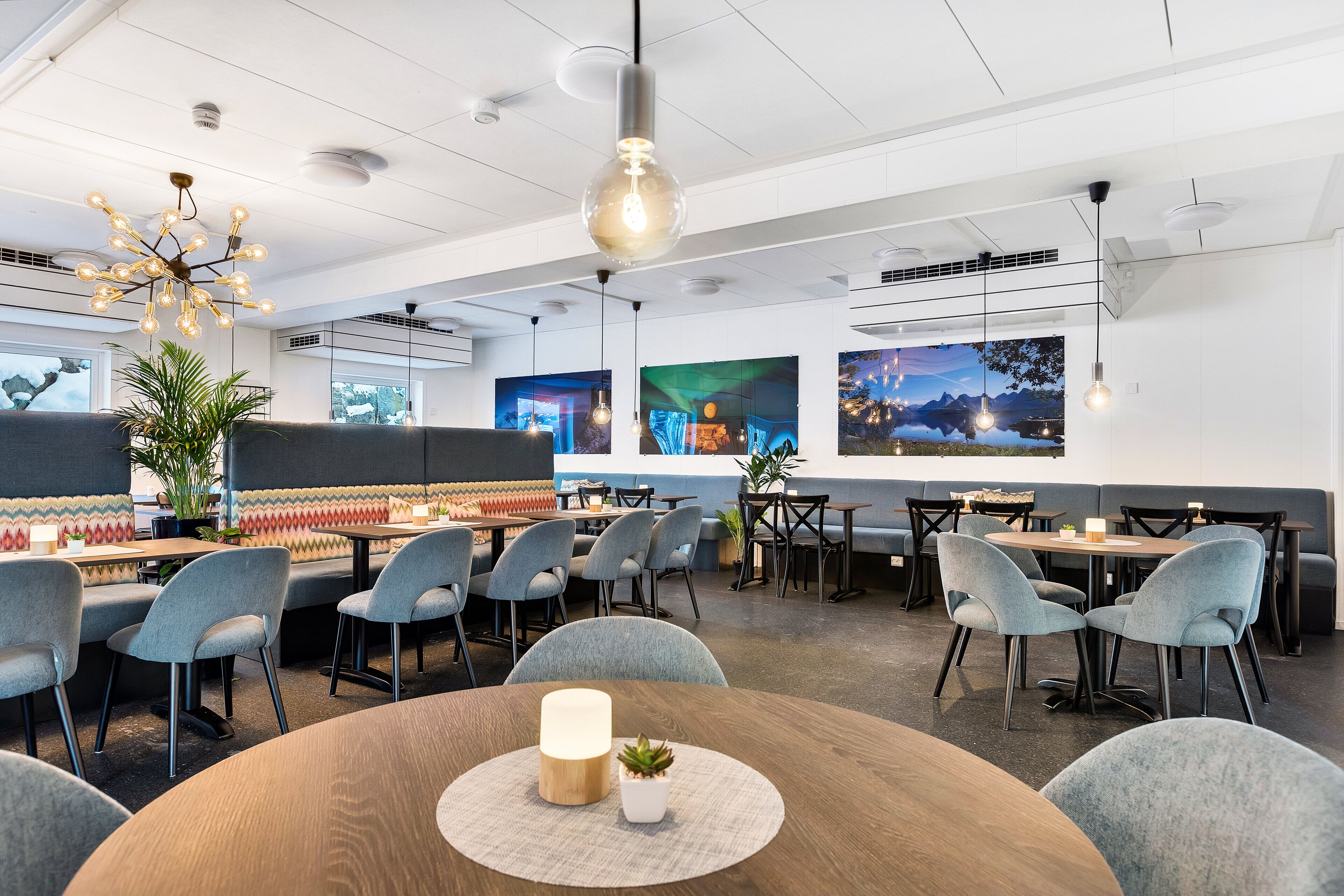 A modern restaurant with round tables, grey upholstered chairs, and a central hanging light fixture.