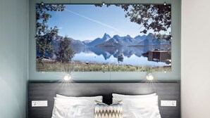 Un dormitorio con cama, almohadas blancas, una almohada colorida, mesitas de noche y un cuadro grande enmarcados con un paisaje de un lago y montañas.