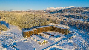 Un hotel grande de varios pisos con un patio central, rodeado por un bosque y montañas cubiertas de nieve.