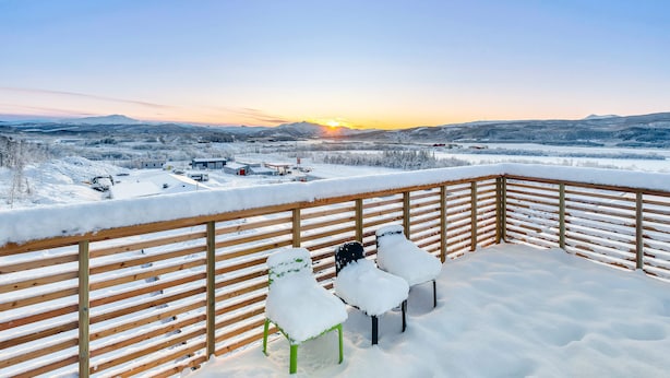 Un muelle cubierto de nieve con dos sillas y barandilla de madera, con vista a un paisaje nevado y una puesta de sol.