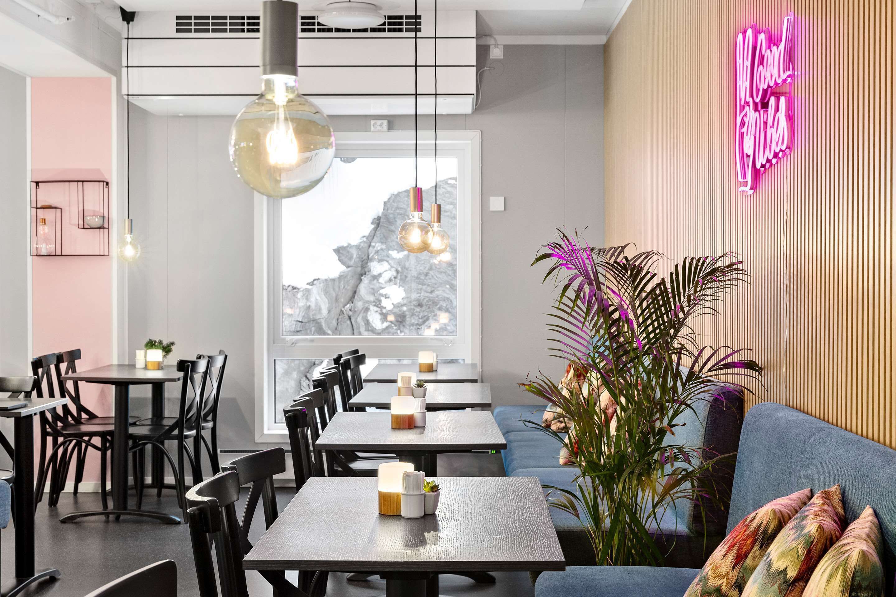 A modern dining area with tables and chairs, a neon sign, and a large window with a mountain view.