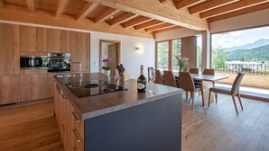 Une cuisine moderne avec des meubles en bois et un grand îlot central, un coin repas avec une table et des chaises, et une vue sur les montagnes grâce à de grandes fenêtres.