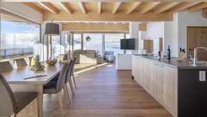 Une cuisine moderne avec des poutres en bois, un coin repas avec une table et des chaises, et une grande fenêtre donnant sur un paysage enneigé.