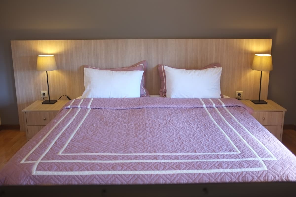A neatly made bed with a pink quilt, two bedside lamps, and a wooden headboard.